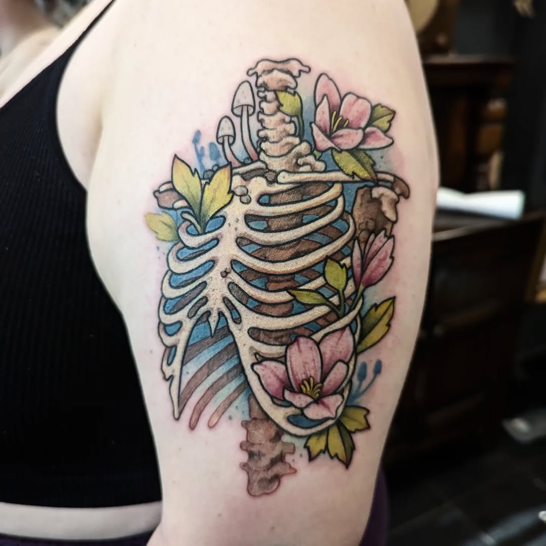 Colored skeletal ribcage and skull tattoo on upper arm with pink flowers, yellow leaves, blue watercolor elements, and mushrooms.