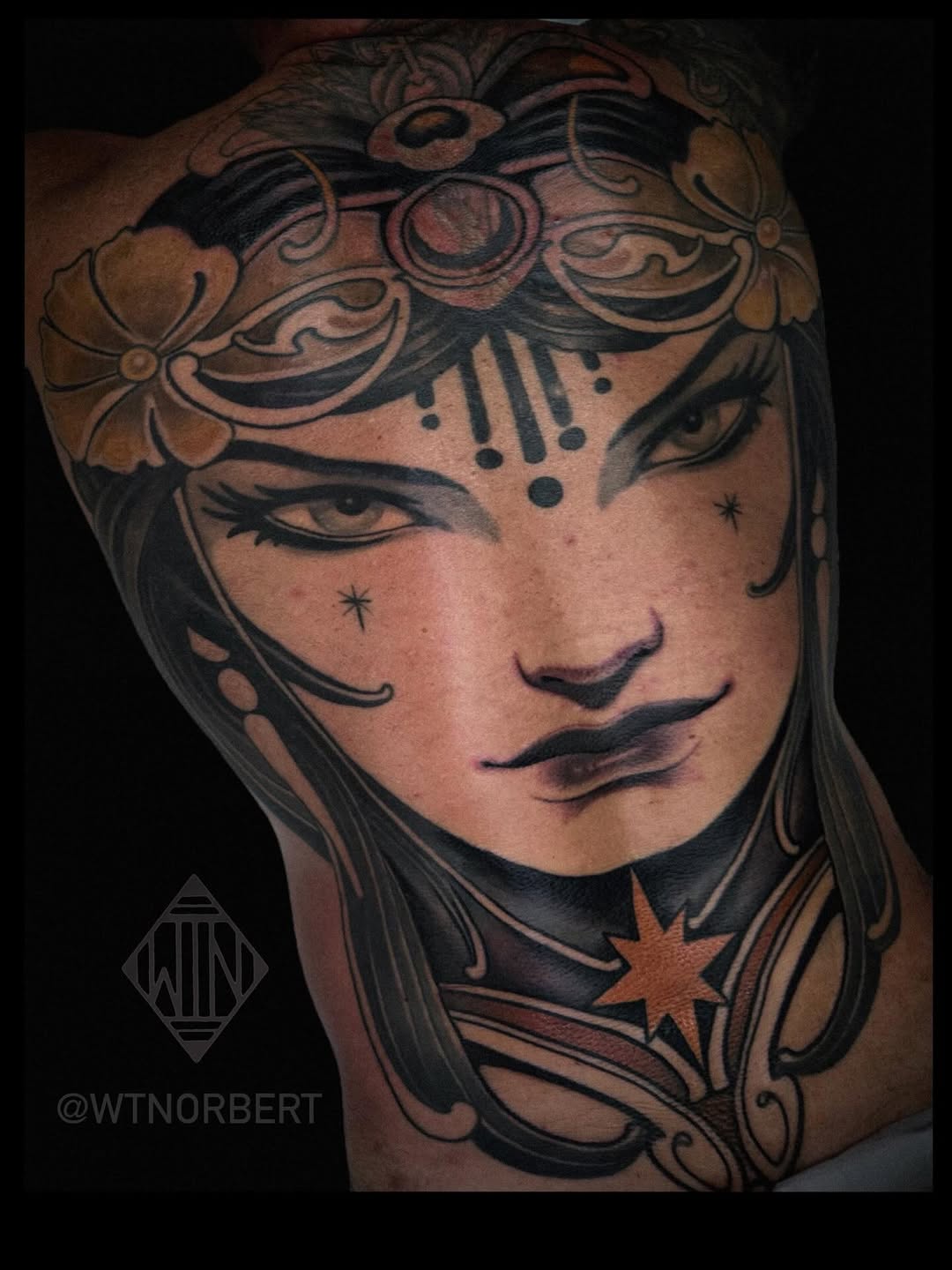 Detailed portrait tattoo of a woman's face with decorative headpiece, floral elements, and star accents in black and brown ink on shoulder.