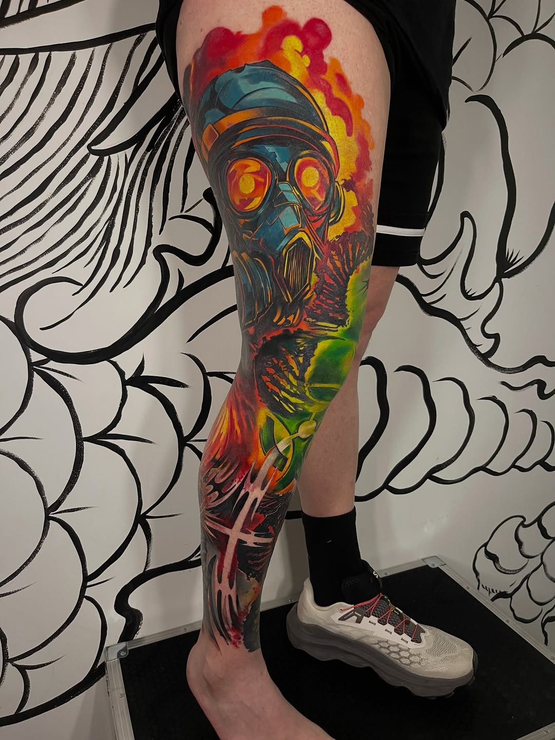 Full-leg sleeve tattoo featuring a skull with glowing eyes and gas mask, surrounded by vibrant red, orange, yellow, and green flames and abstract elements.