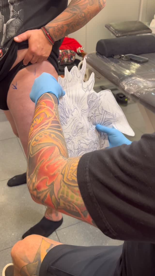 Tattoo artist wearing blue gloves applies a vibrant red and gold flame-patterned design on a client's leg during a studio session.