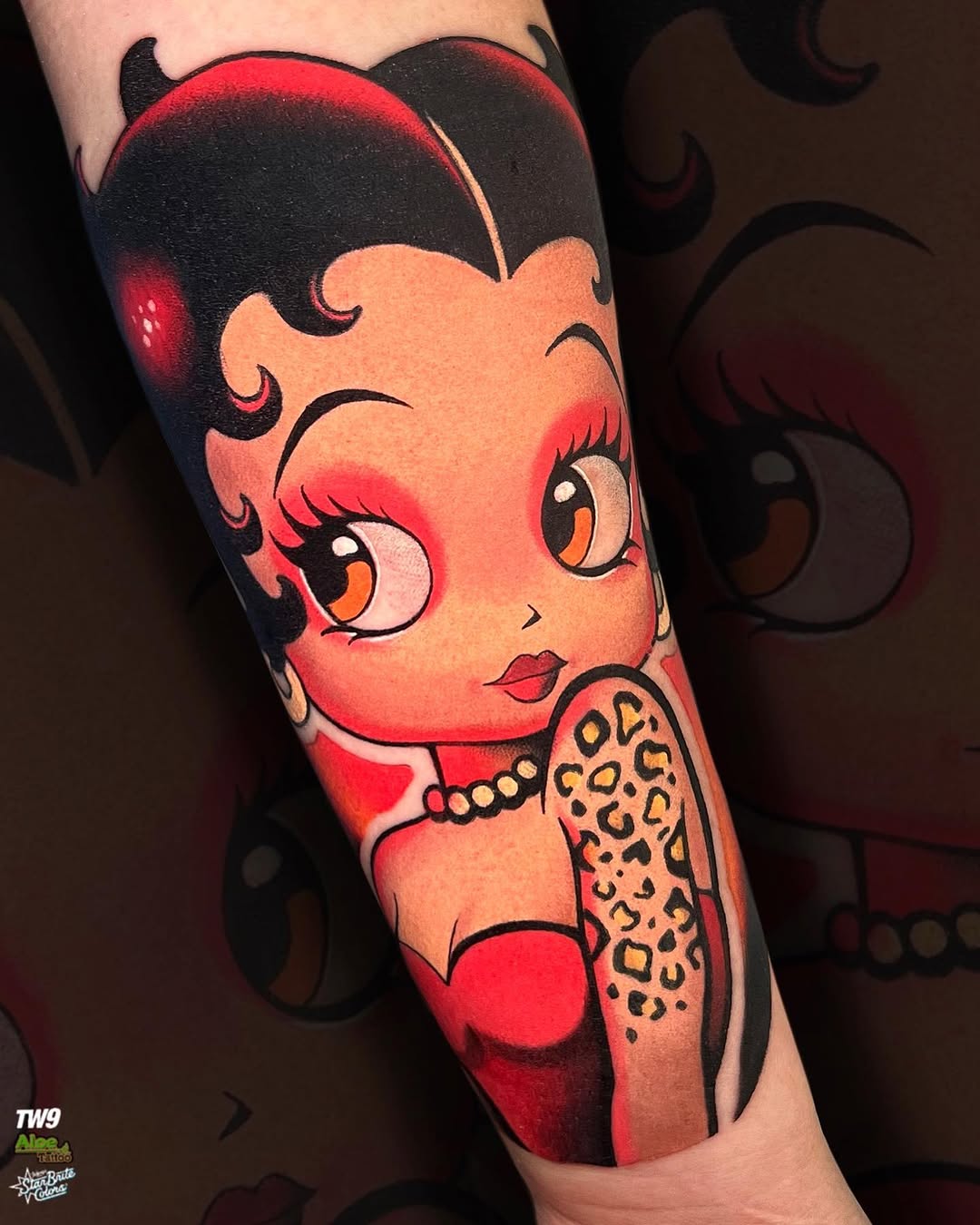 Betty Boop character tattoo on forearm featuring red skin, black hair with horns, large eyes, red lips, pearl necklace, and leopard-print dress against dark background.