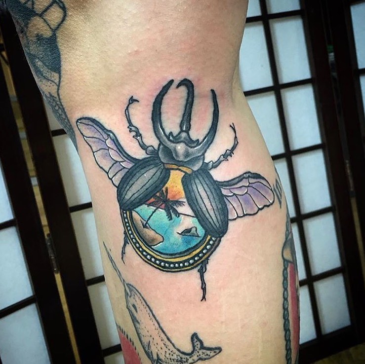 Beetle tattoo with ornate horns, turquoise and gold circular belly detail, translucent wings, and decorative band, placed on the leg.