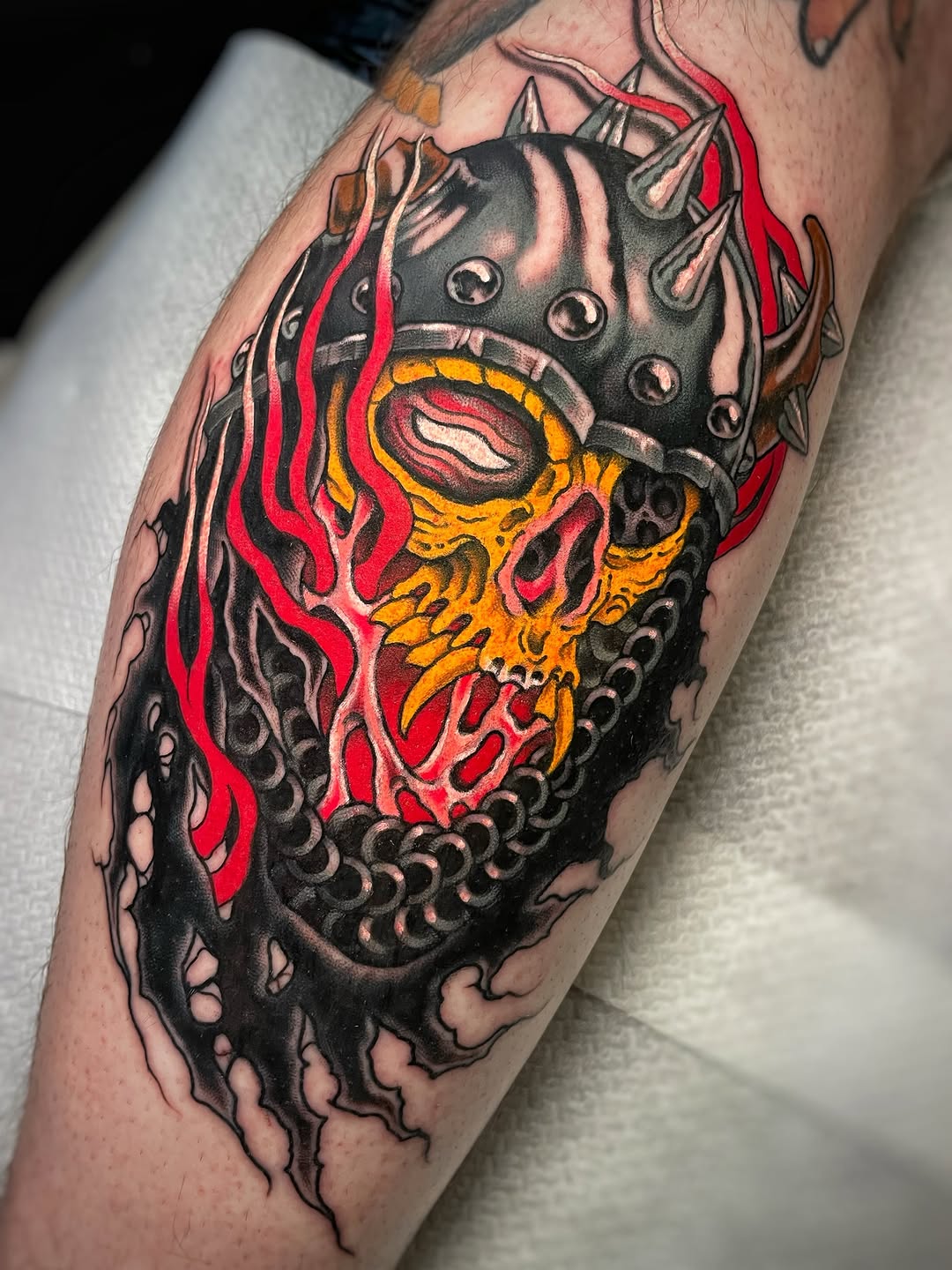 Detailed skull tattoo on calf with red and yellow flames, ornate black scrollwork, spikes, and metallic shading against skin.