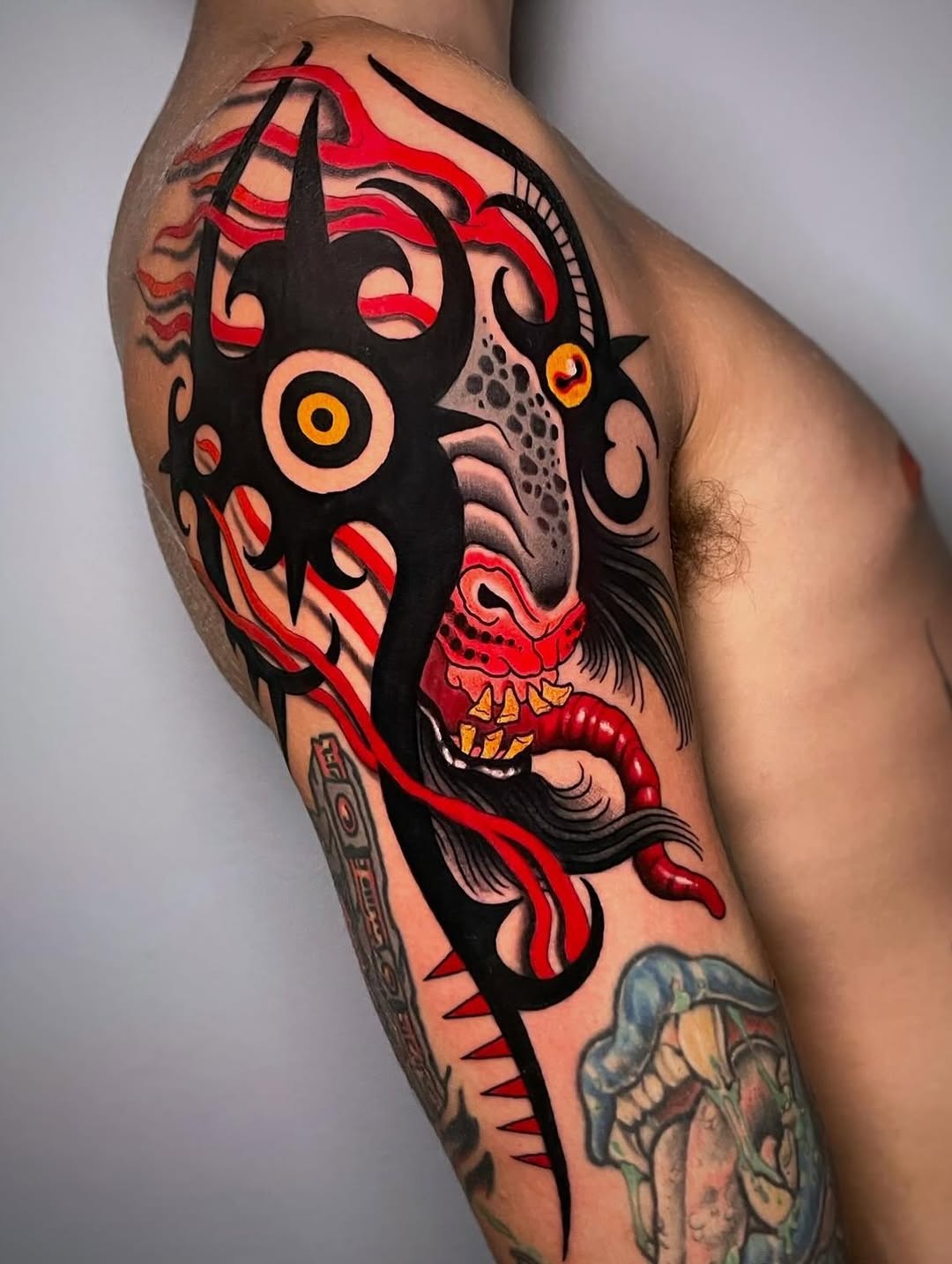 Colorful demon face tattoo on upper arm featuring black silhouette, red flames, golden eyes, and red serpent details against skin.