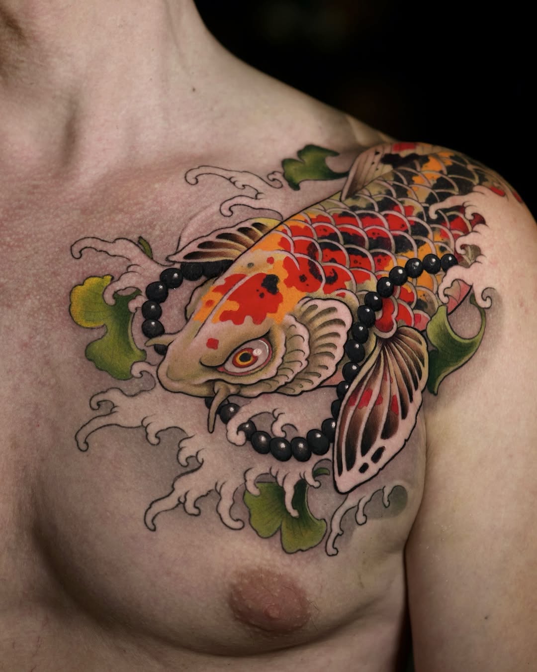 Colorful koi fish tattoo on chest with red, orange, and black scales, green water plants, black beads, and decorative wave elements.