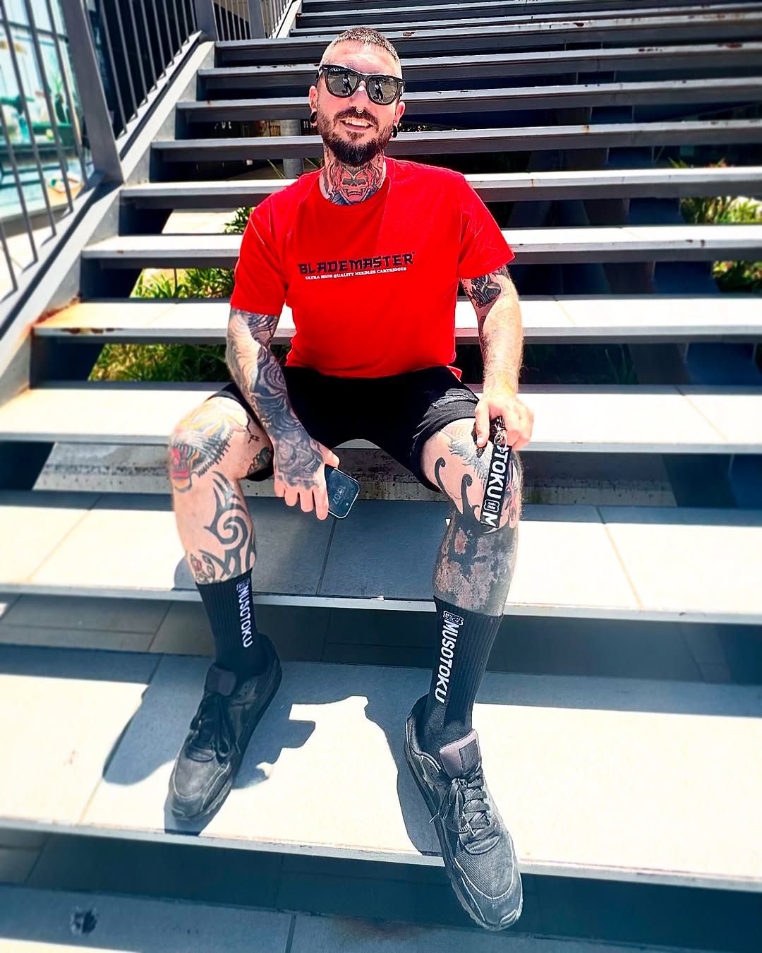 Tattooed man in red shirt sitting on stairs displaying extensive arm and leg tattoos in dark ink across multiple body areas.