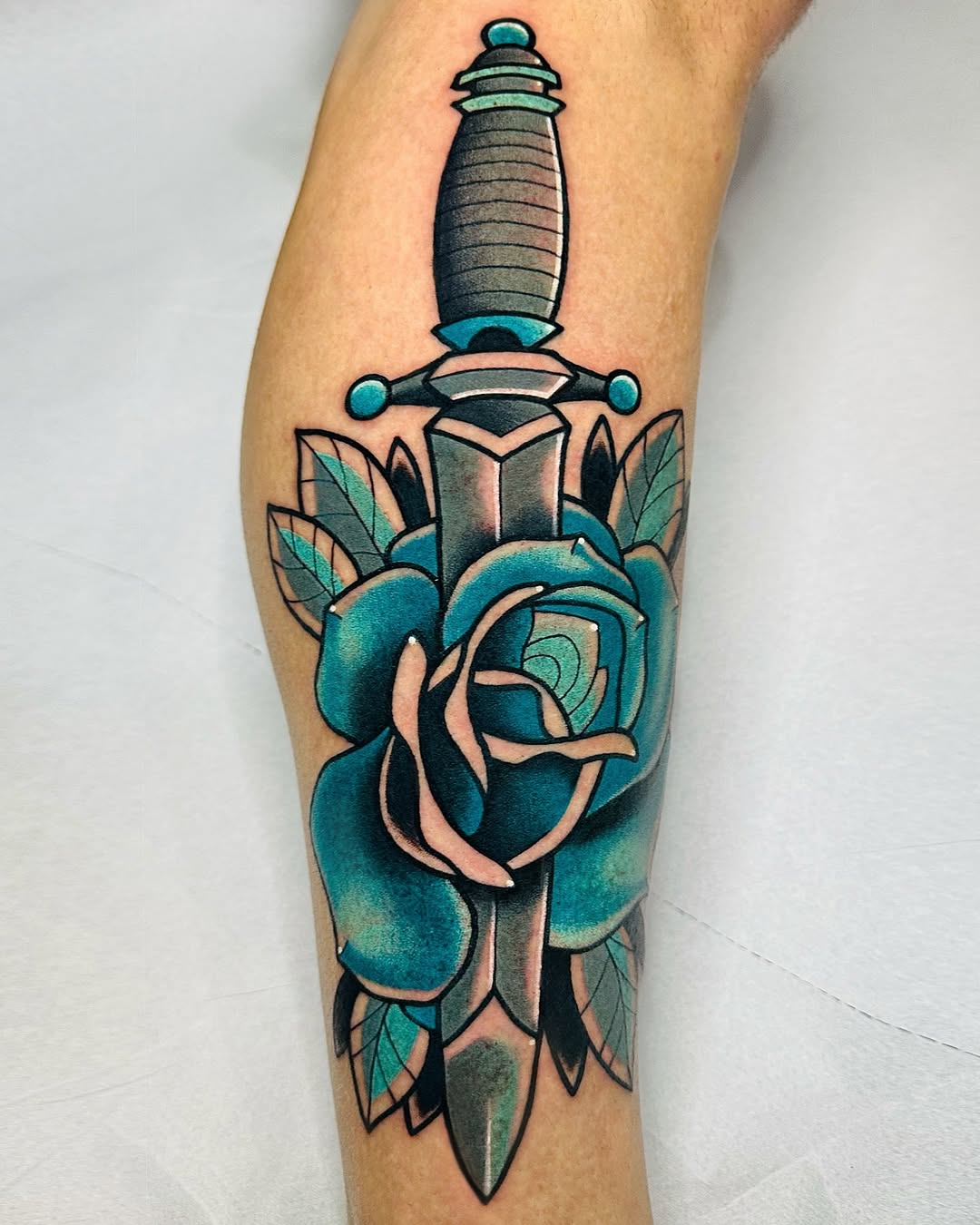 Turquoise rose with green leaves surrounding a dagger with ornamental crossguard, tattooed on the calf in black outline with teal, green, and flesh-tone coloring.