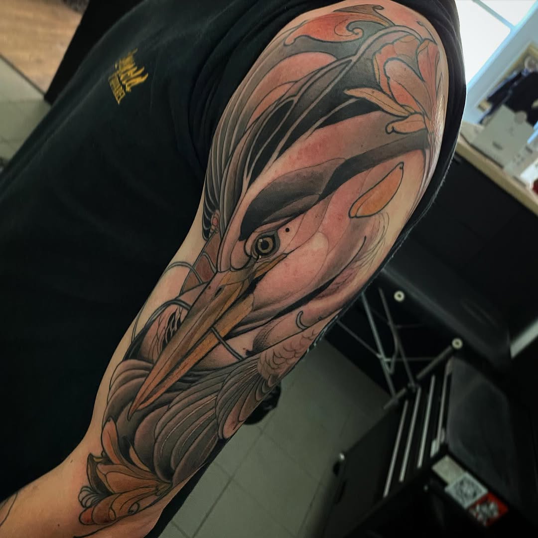 Detailed crane tattoo on the upper arm and shoulder featuring orange, brown, and black ink with ornamental foliage elements.