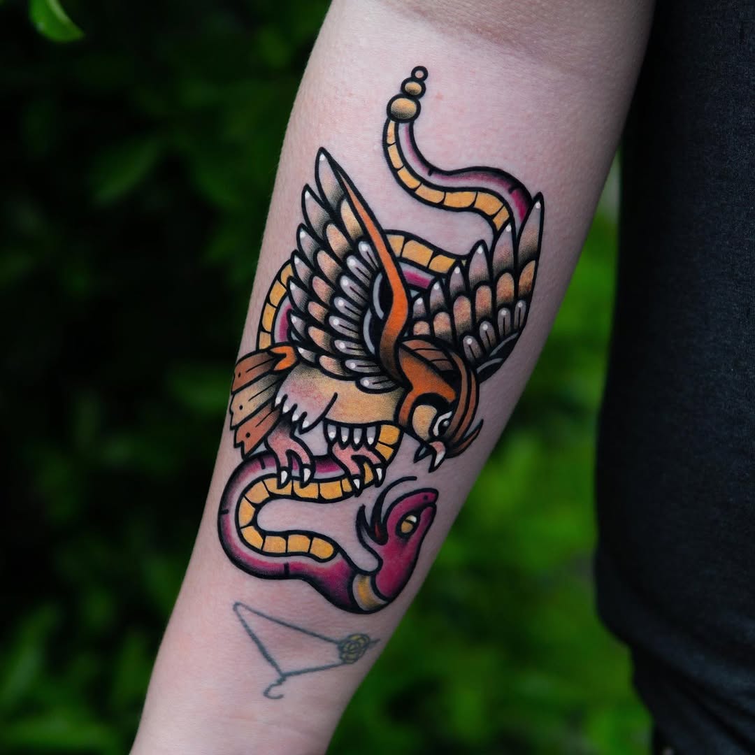 Winged creature with serpents in orange, yellow, pink, and black ink on a forearm, featuring detailed feathered wings and coiled snakes.
