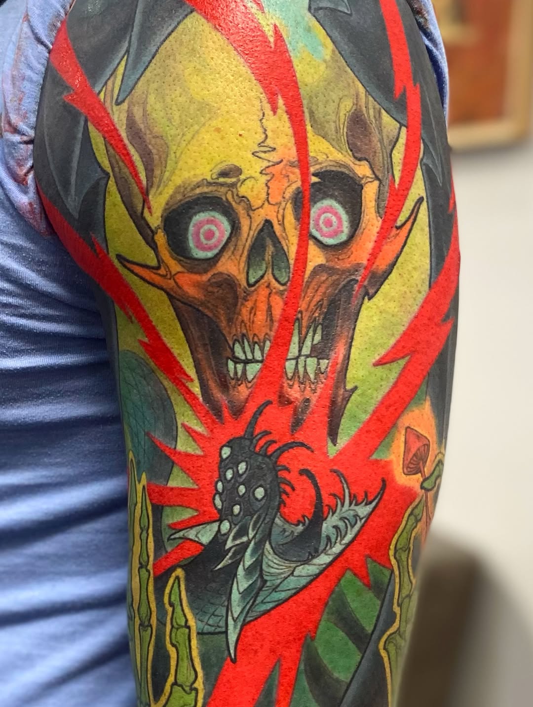 Colorful skull tattoo on upper arm with yellow and green face, red lightning bolts, black eyes with white targets, and a red bird breathing flames below.