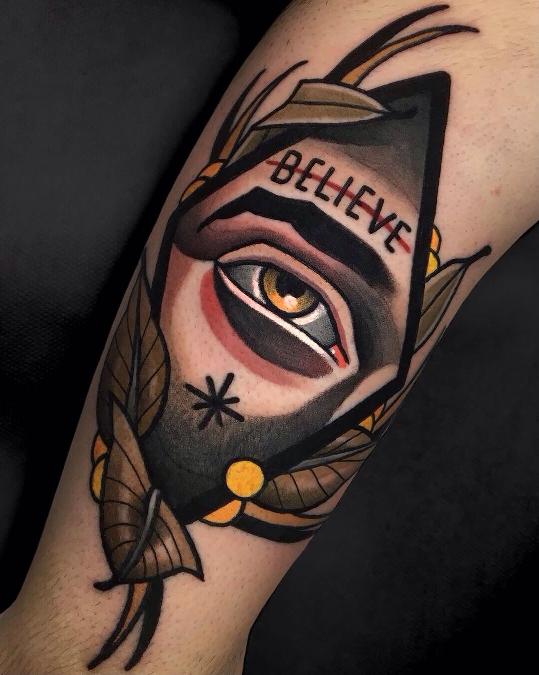 Colorful tattoo on upper arm featuring a geometric cat face with golden eyes, feathered wings, and "BELIEVE" text on the forehead.