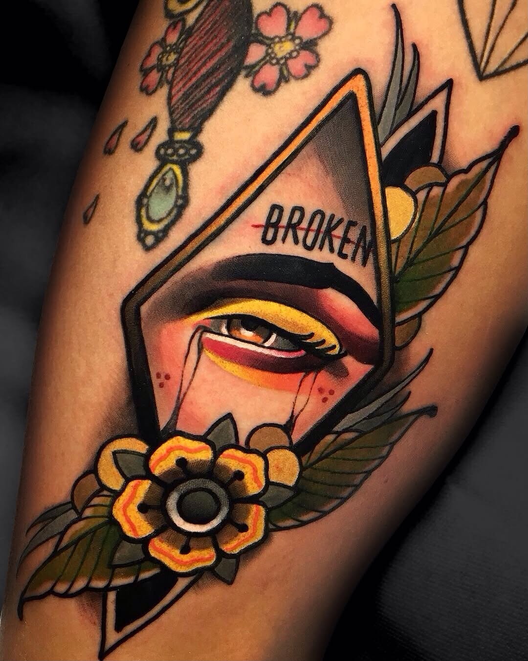 Colorful tattoo on an arm featuring a geometric face with orange and yellow eye makeup, surrounded by flowers and wings, with the word "BROKEN" visible.