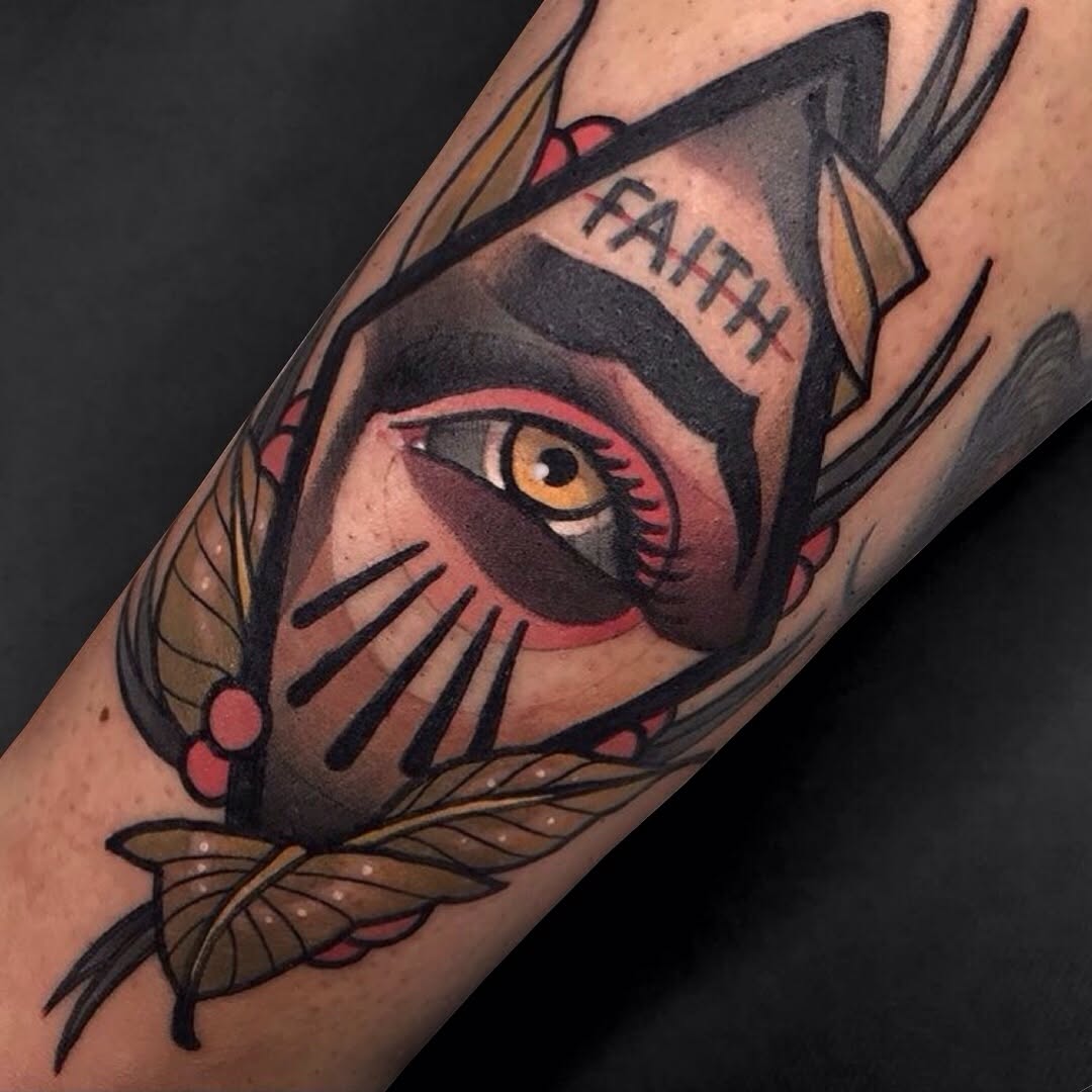 Colored tattoo on forearm featuring a cat face with golden eye, red accents, text reading "FAITH" on forehead, and decorative feather elements in brown and tan tones.
