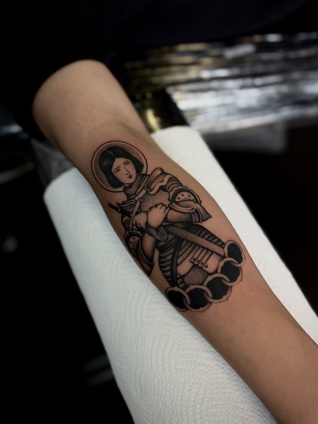 Saint figure with halo holding a sword, seated on clouds, rendered in black ink on the inner forearm.