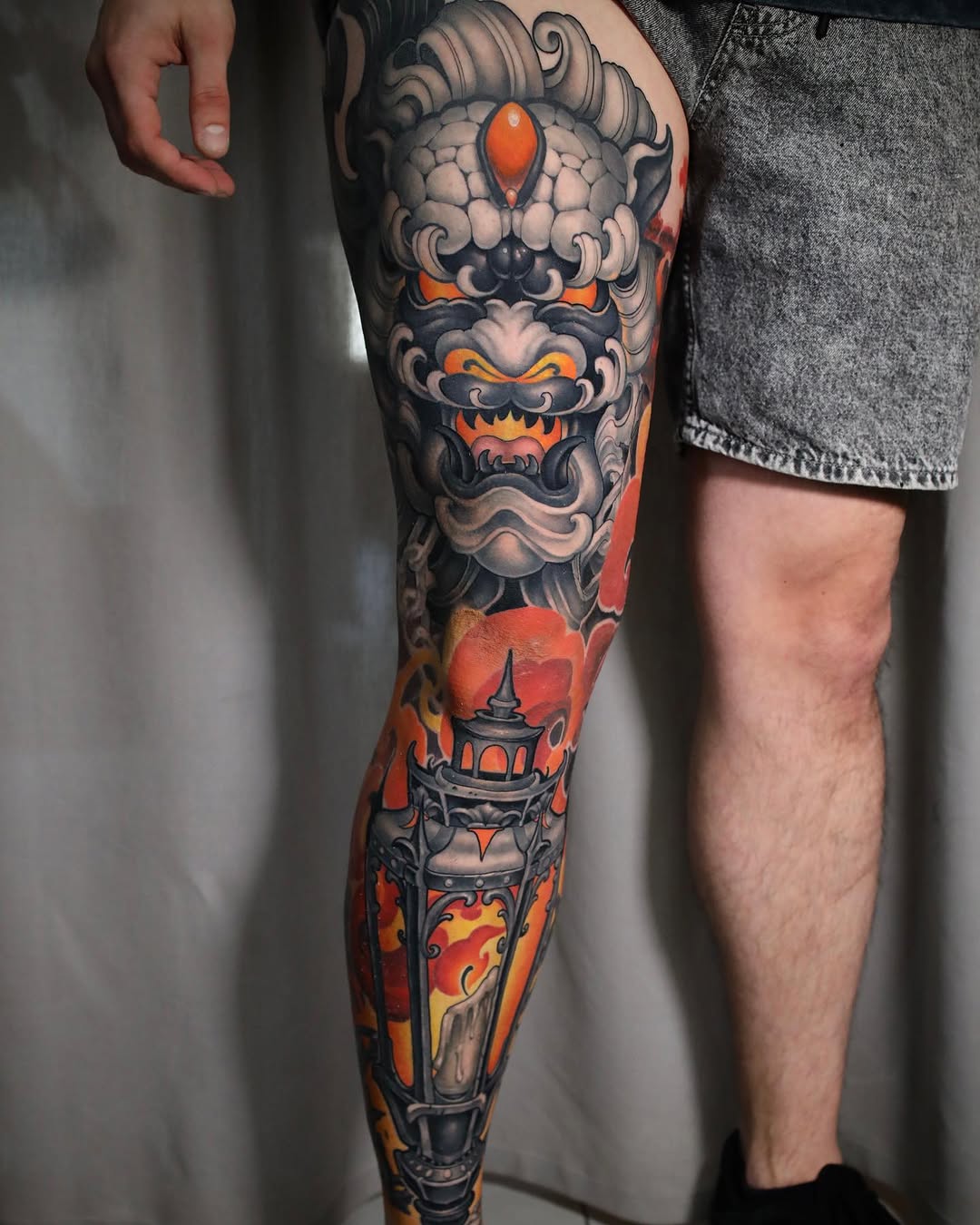 Full-leg tattoo featuring a decorative mask with red gemstone eyes, ornate scrollwork, and a pagoda tower with flames in orange, red, and black ink.