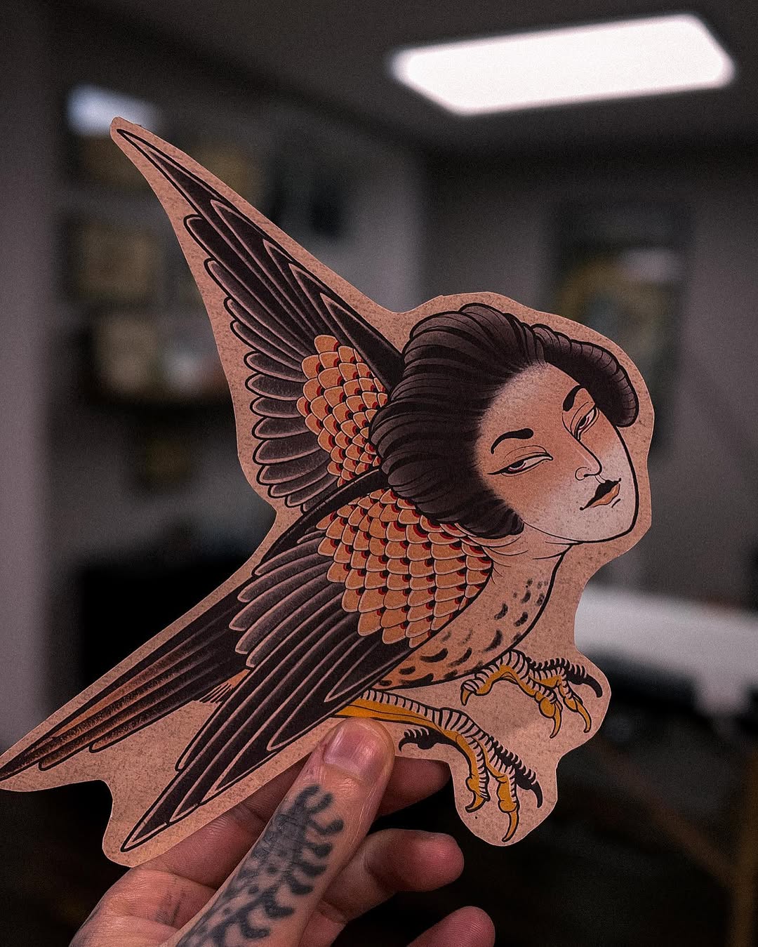 Handheld wooden bird-woman hybrid sculpture with human face, brown hair, orange and black wing details, and yellow taloned feet against a blurred indoor background.