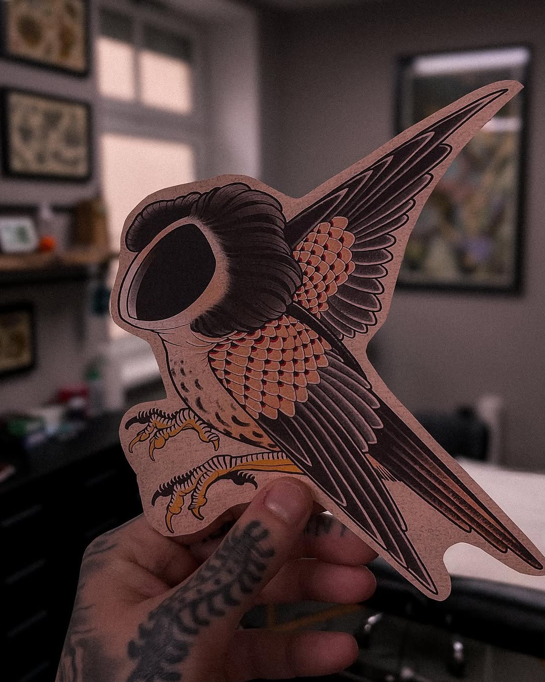 Detailed owl head tattoo design with scaled chest, layered wings, and yellow talons on tan paper, held in tattooed hand indoors.