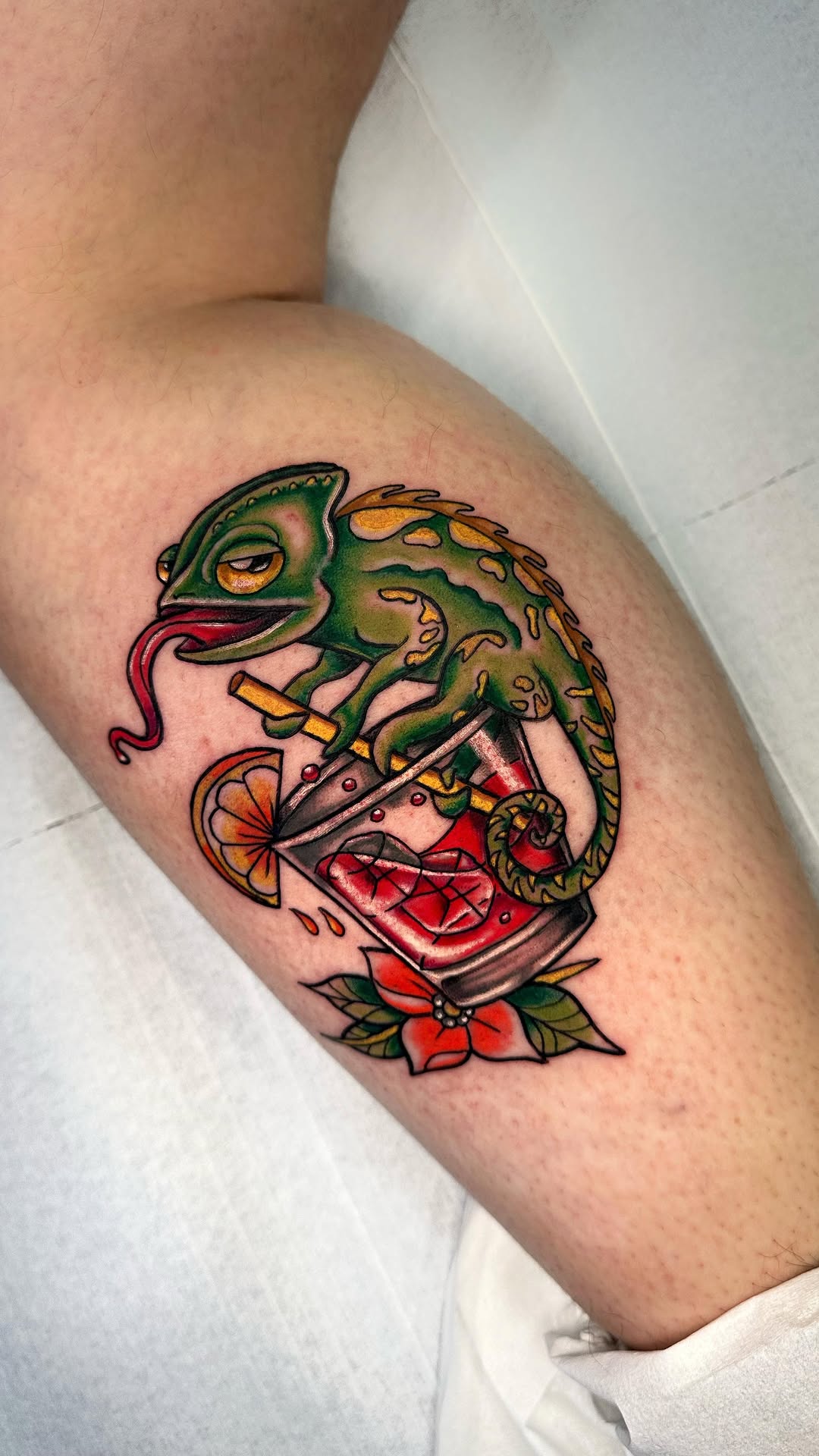 Green chameleon with extended tongue holding an orange slice above a red mouth and flower design, tattooed on the upper leg.